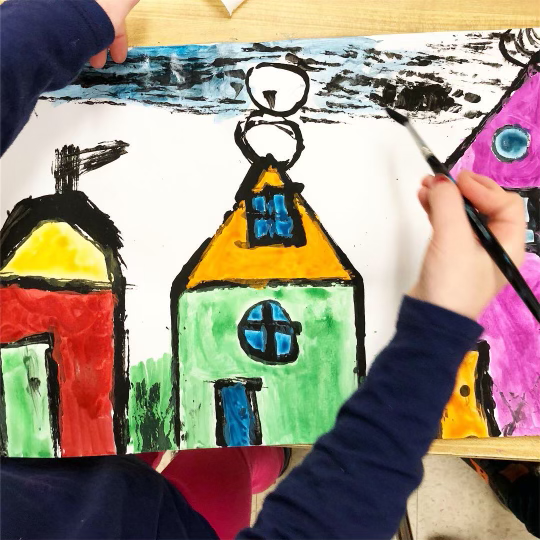 Overhead view of a children's painting.  A child's arm is in the frame holding a paint brush and appears to be actively creating the art.  The artwork depicts several colorful buildings.  