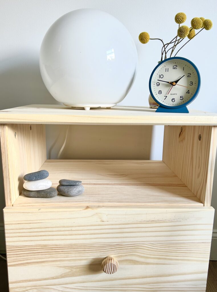 End table from the Hillcrest Psychotherapy office.  The table is unfinished pine wood with an open shelf and drawer.  The shelf includes several small river stones stacked in two piles.  The top of table includes a white spherical lamp, dried flowers, and a analog clock reading eight minutes after nine o'clock.  