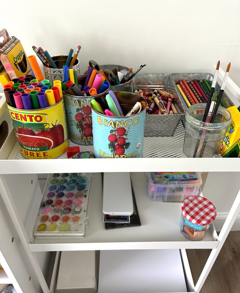 Freestanding storage shelves with art supplies.  Top shelf shows large metal tins containing markers, colored pencils, pencils, crayons, and paint brushes.  Second shelf contains watercolor materials and additional art supplies.  Lowest shelf contains paper.