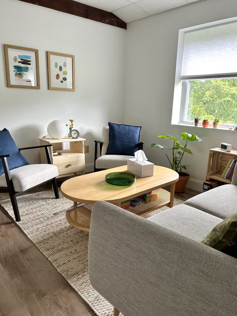 Interior view of the Hillcrest Psychotherapy office.  The foreground shows a gray couch facing a pair of cushioned chairs.  A coffee table, end tables, plants, framed artwork, and colorful throw pillows decorate the room.