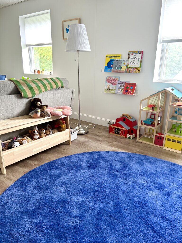 Interior view of children's play space in the Hillcrest Psychotherapy office.  A large, blue, round rug covers the floor.  Books, a dollhouse/ playhouse, stuffed animals, and other therapy toys are visible.  