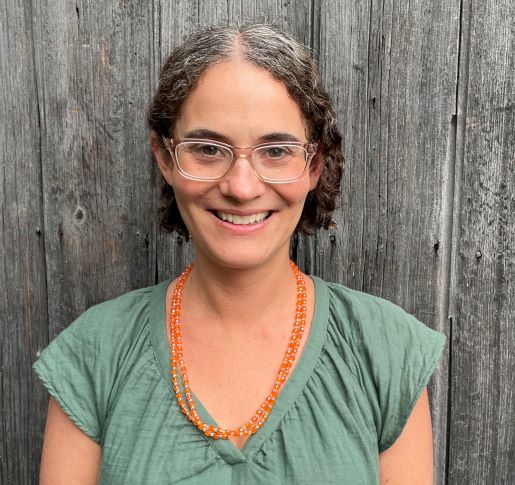 Emily Neuburger smiling with a weathered wood background.  Emily is wearing a light green blouse, orange beaded necklace, and a pair of glasses.  